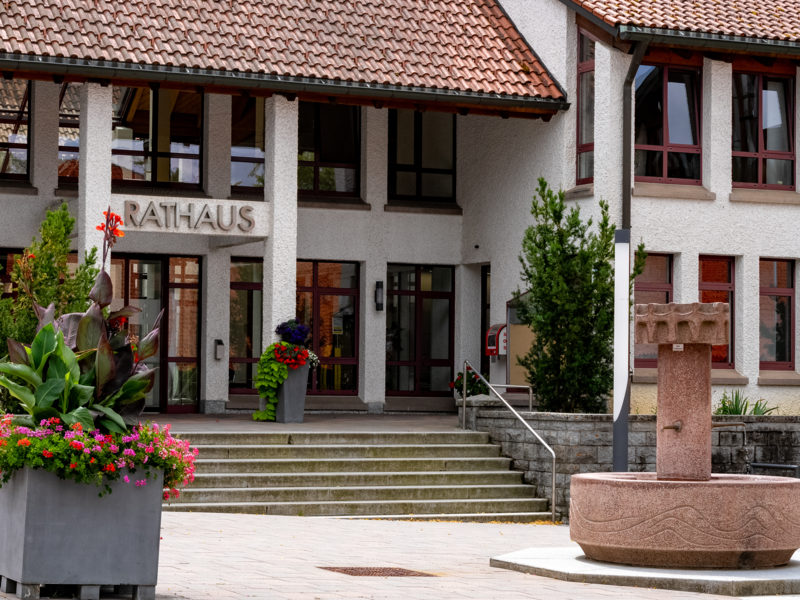 Das Bild zeigt das Brigachaler Rathaus mit einem Vordach, Treppenstufen, einem Springbrunnen und farbenfrohen Blumenkübeln davor.