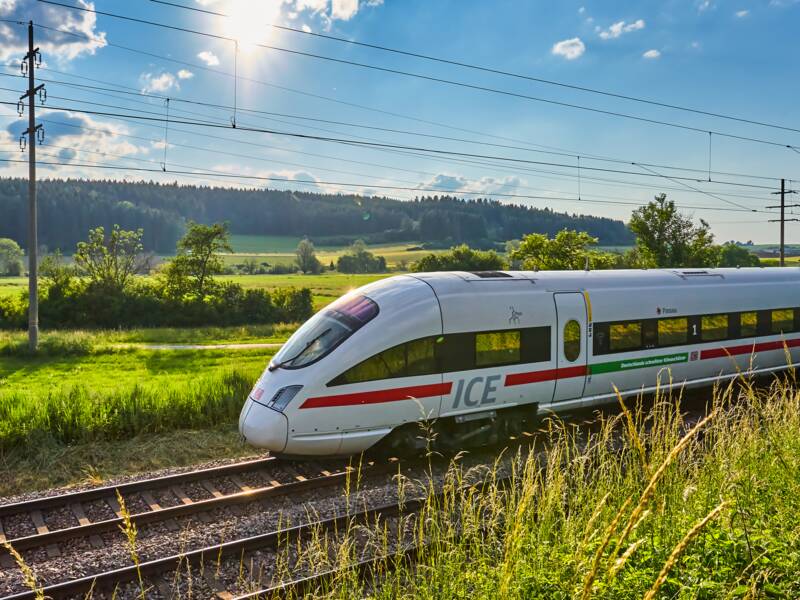 Ein weißer ICE-Zug fährt bei strahlendem Sonnenschein durch eine grüne, ländliche Landschaft.