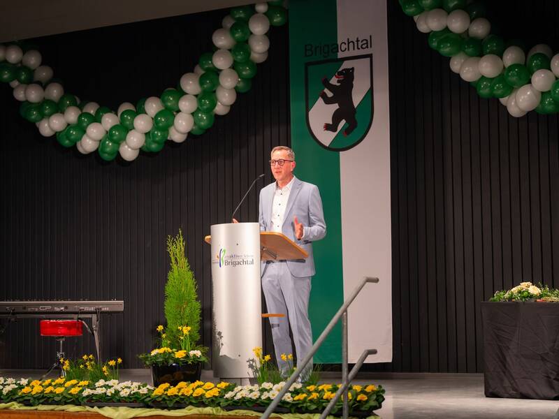 Bürgermeister Michael Schmitt  in einem blauen Anzug spricht auf einer Bühne, dekoriert mit grünen und weißen Luftballons, vor einem Rednerpult mit dem Logo und Schriftzug "Brigachtal".