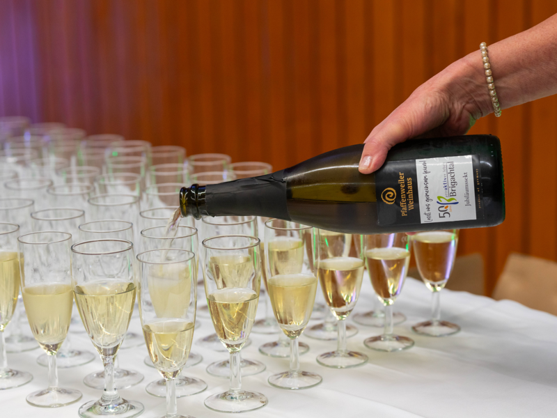 Eine Person gießt Sekt aus einer Flasche in ein Glas, das auf einem Tisch mit vielen gefüllten Gläsern steht.