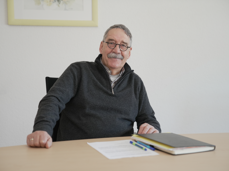 Bruno Braun mit Brille und grauem Pullover sitzt lächelnd an einem Tisch mit Papieren und Stiften.