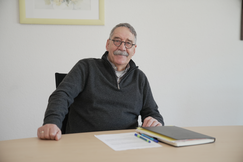 Bruno Braun mit Brille und grauem Pullover sitzt lächelnd an einem Tisch mit Papieren und Stiften.