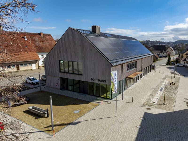 Das Bild zeigt das modernes, rechteckige Dorfhaus mit einem großen Solardach und gepflastertem Vorplatz in einer ländlichen Umgebung.