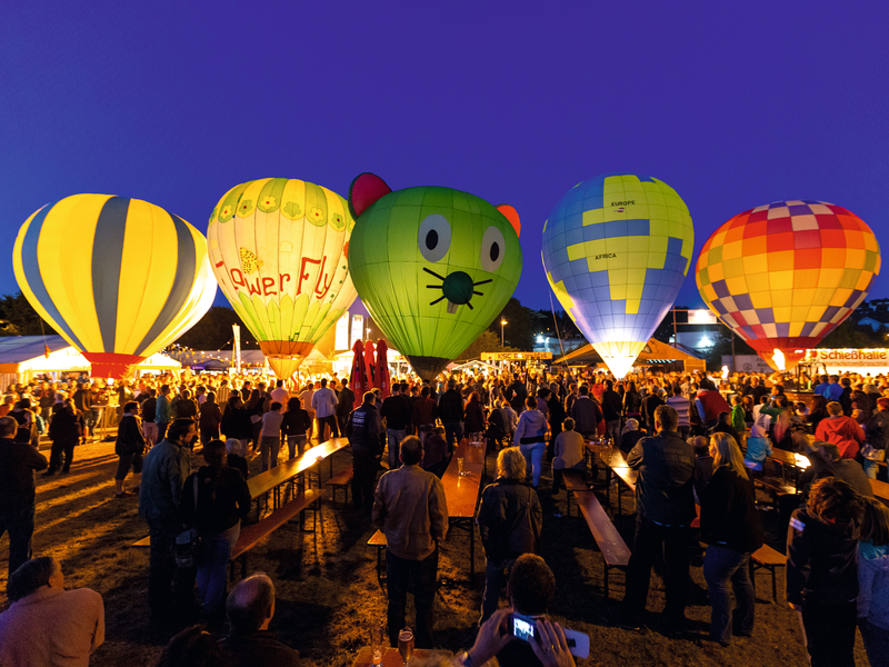 Eine lebhafte Menschenmenge versammelt sich bei Nacht um mehrere bunte Heißluftballons, die über einem Festivalgelände leuchten.