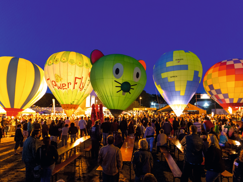 Eine lebhafte Menschenmenge versammelt sich bei Nacht um mehrere bunte Heißluftballons, die über einem Festivalgelände leuchten.