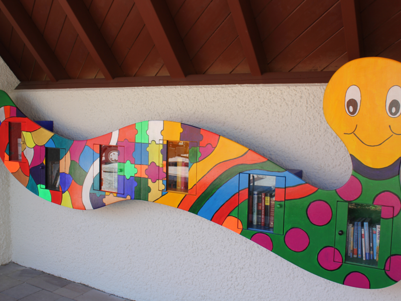 Eine bunte, wellenförmige Bücherwand in Form eines lächelnden Tausendfüßlers mit mehreren kleinen Regalen enthält Bücher.