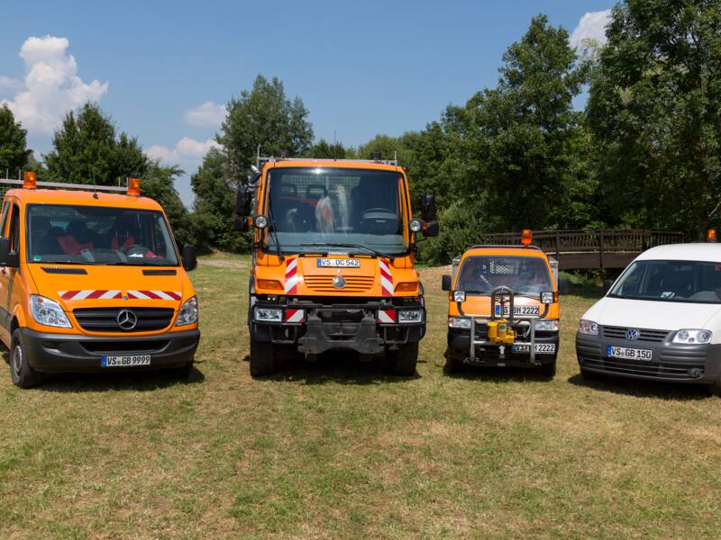 Vier Fahrzeuge, darunter drei orange und ein weißes, stehen auf einer Wiese vor einem Hintergrund mit Bäumen und einem blauen Himmel.