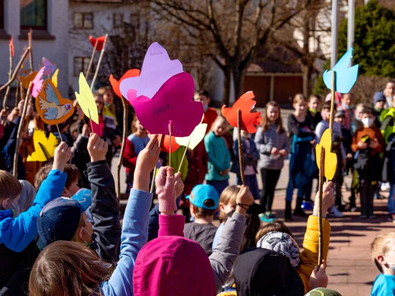Eine Gruppe von Kindern hält bunte Papierfiguren an Stöcken in die Höhe, während sie draußen zusammenstehen.