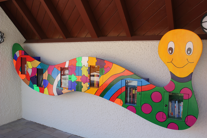 An einer weißen Wand ist ein bunter Bücherregal in Form einer fröhlichen, lächelnden Raupe montiert.