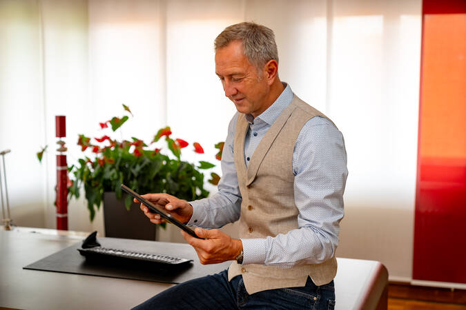 Bürgermeister Michael Schmitt  in einem Büro liest ein Tablet und trägt ein hellblaues Hemd und eine beige Weste, während im Hintergrund rote Blumen zu sehen sind.