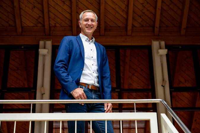 Bürgermeister Michael Schmitt in einem blauen Sakko steht lächelnd auf einem hellen Treppenabsatz vor einer hölzernen Dachkonstruktion.