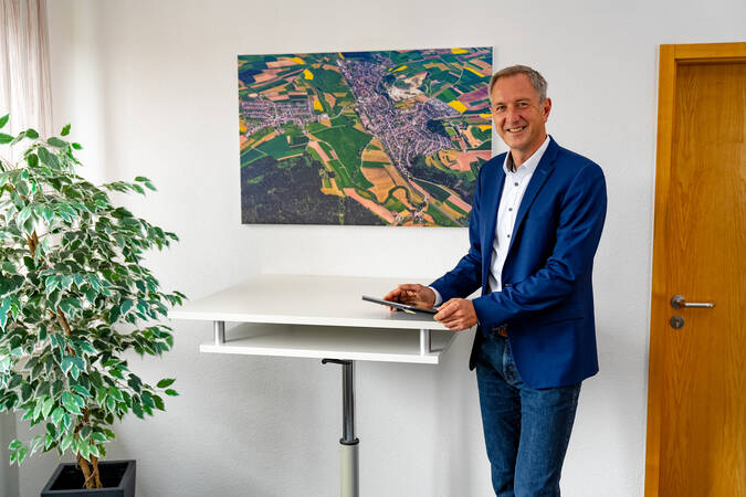 Bürgermeister Michael Schmitt in einem blauen Sakko steht lächelnd an einem Stehtisch in einem Büro neben einer Pflanze und hinter ihm hängt ein Luftbild von Brigachtal an der Wand.