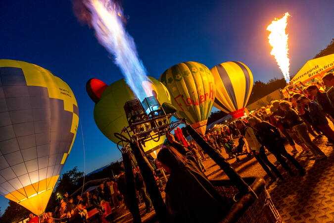 Auf einem abendlichen Fest steigen mehrere bunte Heißluftballons mit brennenden Flammen in den Himmel, während eine Gruppe von Menschen zuschaut.