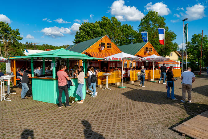 Mehrere Menschen genießen einen sonnigen Tag auf einem festlichen Platz beim Dorffest mit bunten Verkaufsständen und überdachten Sitzbereichen.