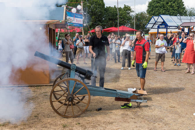 Beim Dorffest beobachtet eine Gruppe von Menschen, wie Bürgermeister Michael Schmitt eine historische Kanone abgefeuert wird, während Rauch aufsteigt.