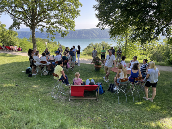 Eine Gruppe von Menschen sitzt und steht im Schatten von Bäumen auf einer Wiese mit Blick auf einen See in der Ferne.