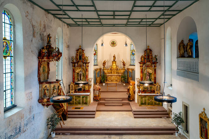 Der helle Innenraum einer Kirche zeigt einen aufwendig gestalteten Altar mit mehreren Heiligenstatuen und verzierten Fenstern.