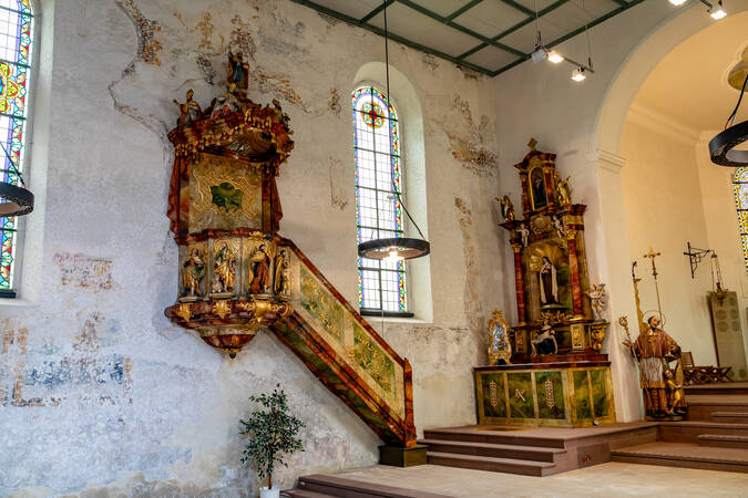 Eine historische Kirche mit einer verzierten Kanzel und einem kunstvollen Altar vor bunt verglasten Fenstern.