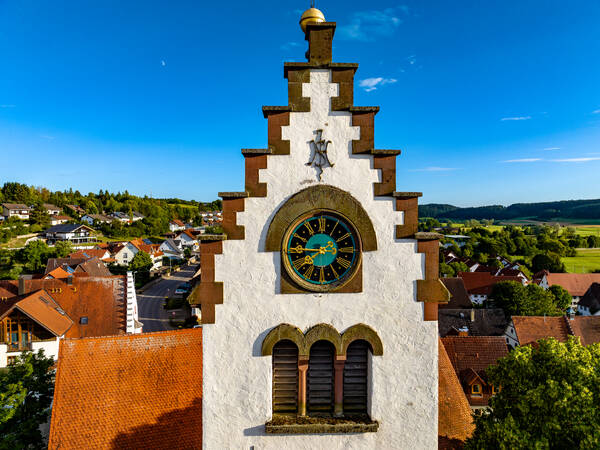 Eine weiße Kirchturmuhr der Kirche St. Martin mit römischen Ziffern thront über einer malerischen Dorflandschaft mit roten Dächern und bewaldeten Hügeln im Hintergrund.