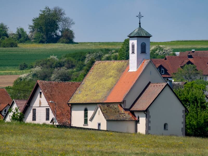 Eine ländliche Kirche mit einem weißen Turm und roten Dächern steht vor einer grünen Hügellandschaft und weiteren Häusern.