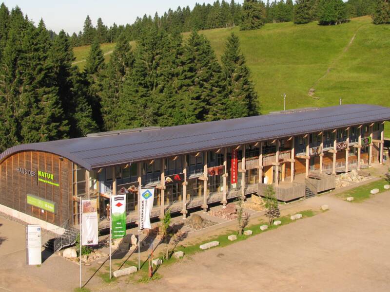 Ein langes Gebäude mit Solardach steht am Waldrand, umgeben von grünen Wiesen und hohen Bäumen.