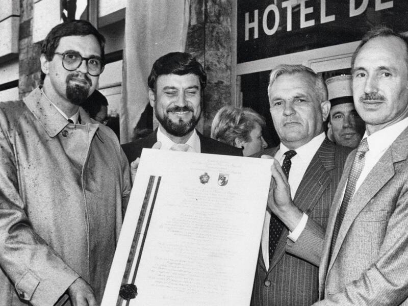 Vier Männer in Anzügen halten ein großes Dokument vor einem Gebäude mit der Aufschrift "HOTEL DE VILLE".