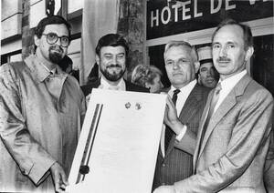 Vier Männer in Anzügen halten ein großes Dokument vor einem Gebäude mit der Aufschrift "HOTEL DE VILLE".