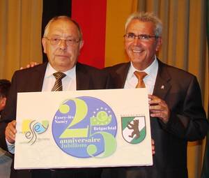 Zwei Männer in Anzügen halten ein Schild mit der Aufschrift "25ème anniversaire Jubiläum", das die Partnerschaft zwischen Essey-lès-Nancy und Brigachtal feiert.