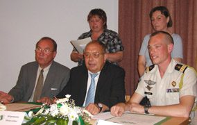 Drei Männer sitzen an einem Tisch mit Blumenarrangement, während zwei Frauen stehend im Hintergrund zusehen.