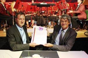 Bürgermeister Michael Schmitt und Bürgermeister Michel Breuille sitzen am Tisch und halten gemeinsam ein Dokument, während im Hintergrund ein Orchester in roten Uniformen spielt.