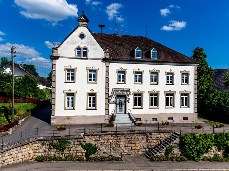 Ein zweistöckiges, historisches Gebäude mit symmetrischen Fenstern und einem Satteldach steht vor einem klaren blauen Himmel, umgeben von gepflegter Vegetation.