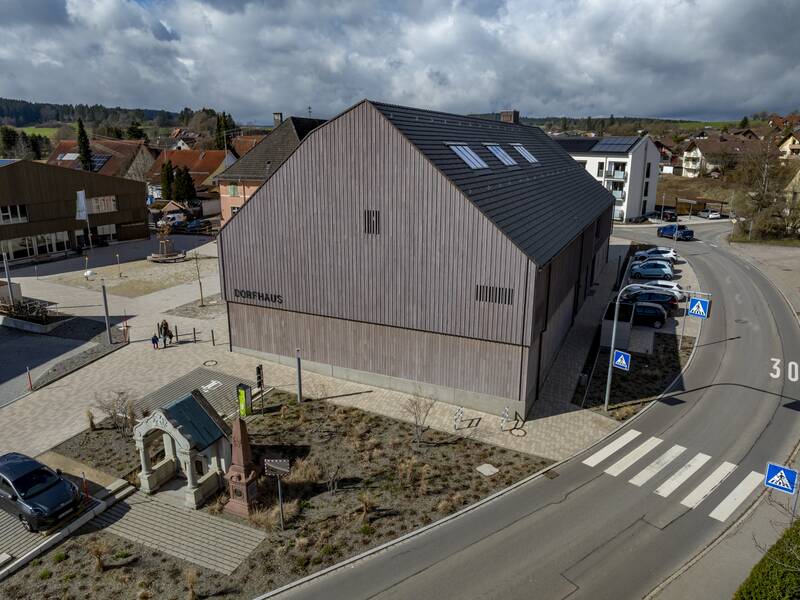 Ein modernes Gebäude mit der Aufschrift "Dorfhaus" steht an einer Kreuzung mit Zebrastreifen und parkenden Autos, umgeben von einem Platz und weiteren Gebäuden im Hintergrund.