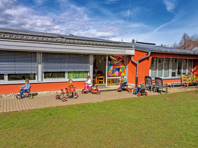 Vor einem flachen, orangefarbenen Gebäude der Kita Gaisberg mit großen Fenstern spielen mehrere Kinder auf Dreirädern auf einem Gehweg.