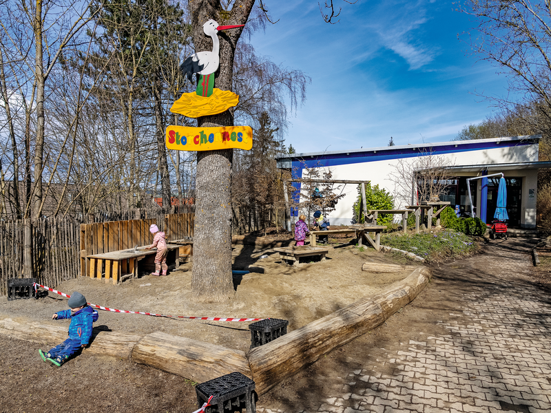 Ein Spielplatz mit Holzbänken, einem Baum mit Beschriftung und spielenden Kindern vor der Kita Storchennest.