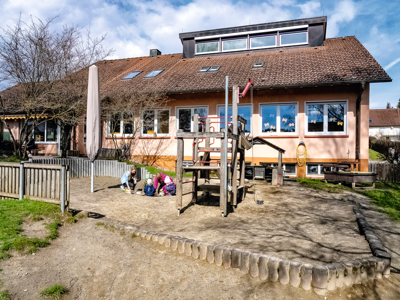 Ein Spielplatz mit Holzstruktur und drei spielenden Kindern befindet sich vor einem Gebäude  der Kita am Vorberg mit großen Fenstern und rotem Dach.
