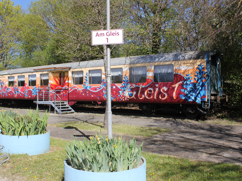 Ein alter Eisenbahnwaggon, bunt mit Graffiti bemalt, steht auf einem Platz mit einem Schild "Am Gleis 1" in einer grünen Umgebung.