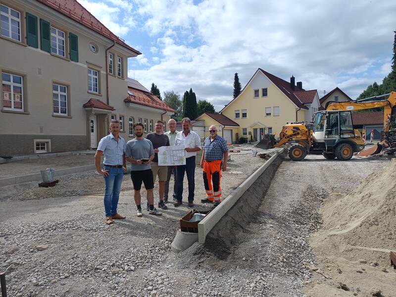 Eine Gruppe von sechs Personen steht auf einer Baustelle vor einem Gebäude, während sie Baupläne betrachten.