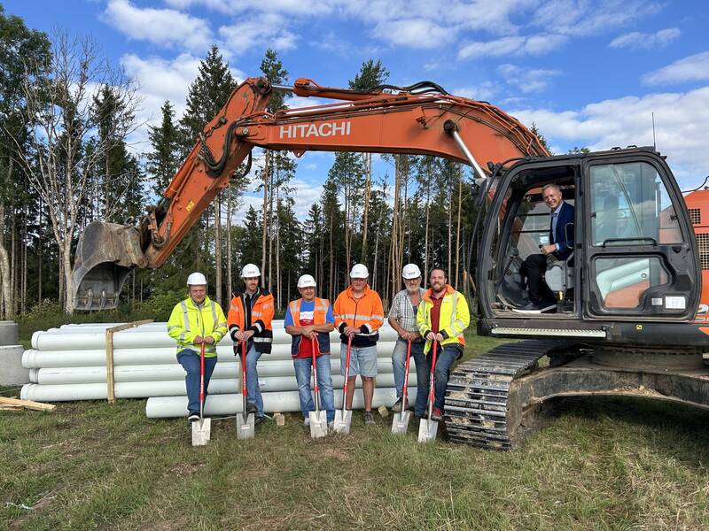 Sechs Personen in Arbeitskleidung und Helmen posieren mit Schaufeln vor einem großen Bagger, umgeben von Rohren und Wald im Hintergrund.