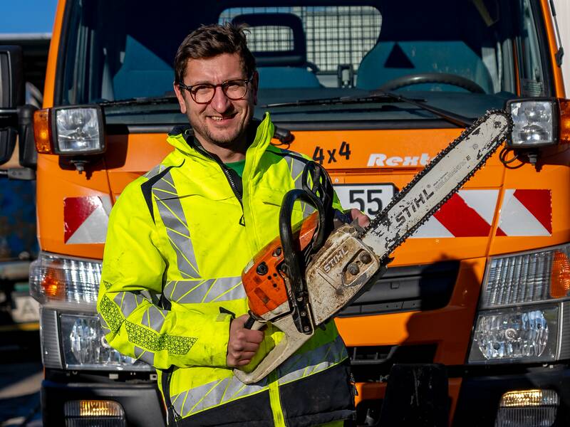Valentin Klemann in einer gelben Warnjacke steht vor einem orangefarbenen Fahrzeug und hält eine Kettensäge.