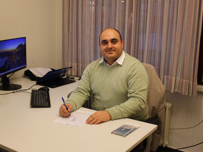 Khaled Mousa sitzt an einem Schreibtisch in einem Büro, trägt einen grünen Pullover und schreibt auf ein Dokument, während er lächelnd in die Kamera schaut.