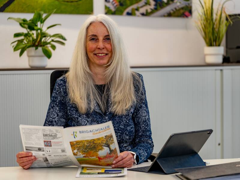 Sandra Sowinski sitzt lächelend an einem Tisch, hält ein Mitteilungsblatt und hat ein Tablet vor sich.