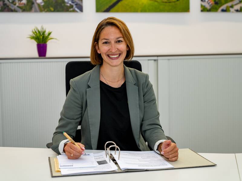 Christine Schafbuch in einem grauen Blazer sitzt lächelnd an einem Schreibtisch mit Akten und einem Stift in der Hand, im Hintergrund sind Pflanzen und Bilder an der Wand zu sehen.