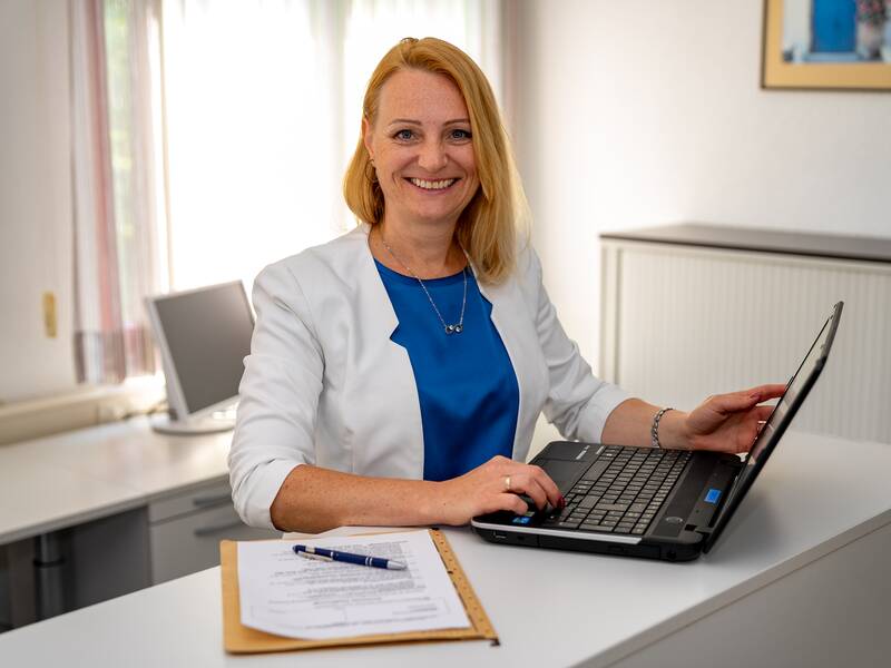 Marina Ritscher sitzt lächelnd an einem Schreibtisch mit einem Laptop, einem Klemmbrett und einer Büroeinrichtung im Hintergrund.