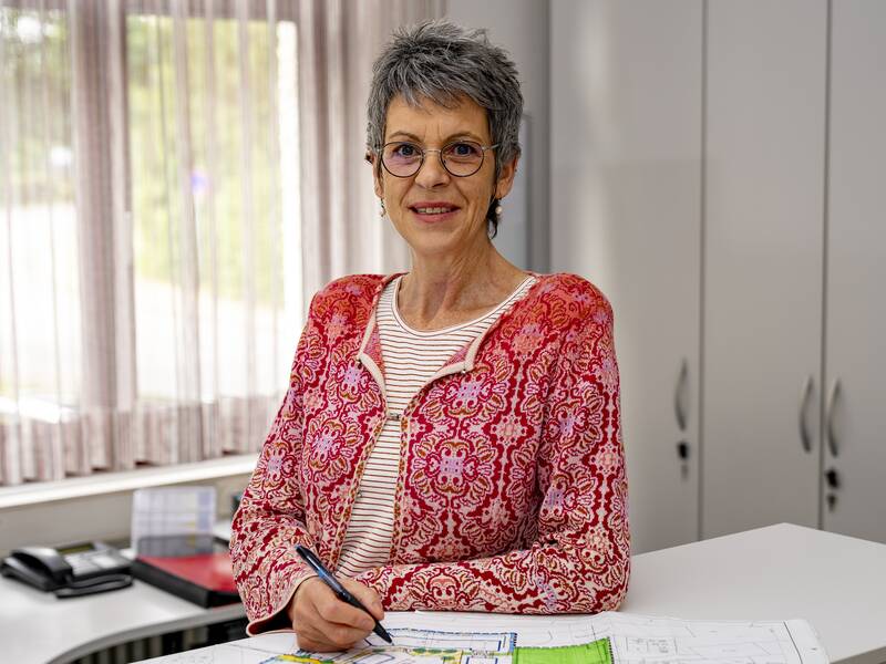 Annette Albiez mit Brille und gemustertem Oberteil steht an einer Theke und hält einen Stift, während vor ihr ein Plan liegt.