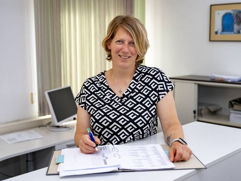 Sandra Dold steht lächelnd an einem Tresen und hält einen Stift, umgeben von Bürogegenständen und Unterlagen.