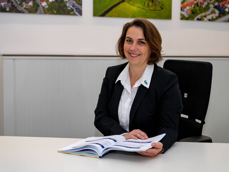 Martina Enderlein sitzt an einem Tisch und hält ein geöffnetes Buch in der Hand, während sie in die Kamera lächelt.