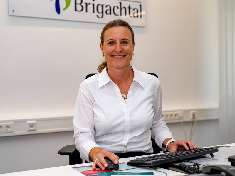 Nicole Perkuhn sitzt lächelns in Büroumgebung an einem Schreibtisch mit Computer.
