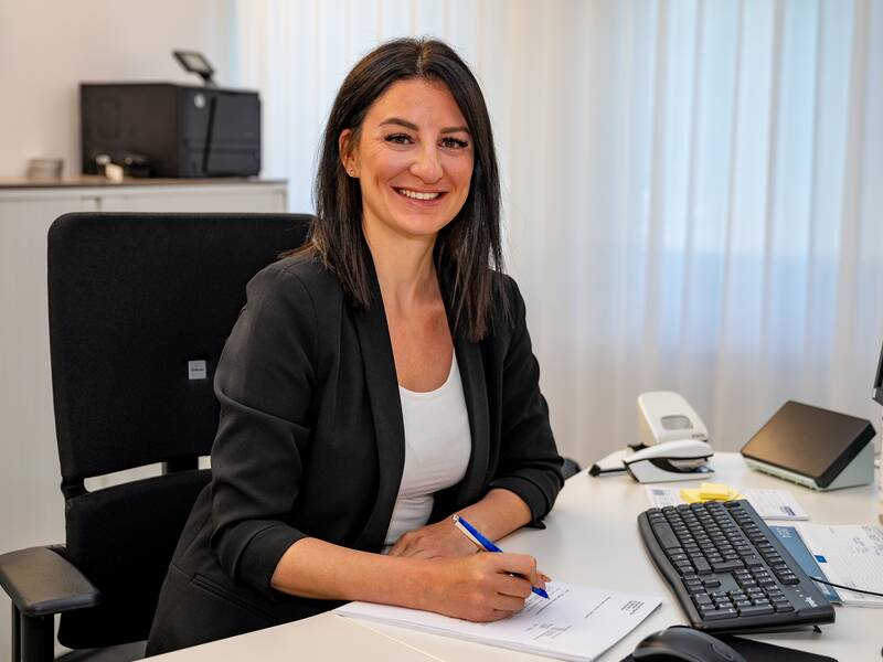 Denise Stöcker sitzt lächelnd an einem Schreibtisch und hält einen Stift in der Hand, umgeben von Büroausstattung.