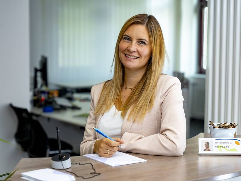 Sandra Zipfel sthet lächelend in einem Büro und hält einen Stift in der Hand.