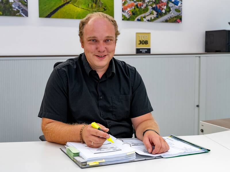 Eric Elsäßer sitzt lächelnd an einem Tisch in einem Büro und hält einen gelben Textmarker in der Hand, während er sich über ein aufgeschlagenes Dokument beugt.
